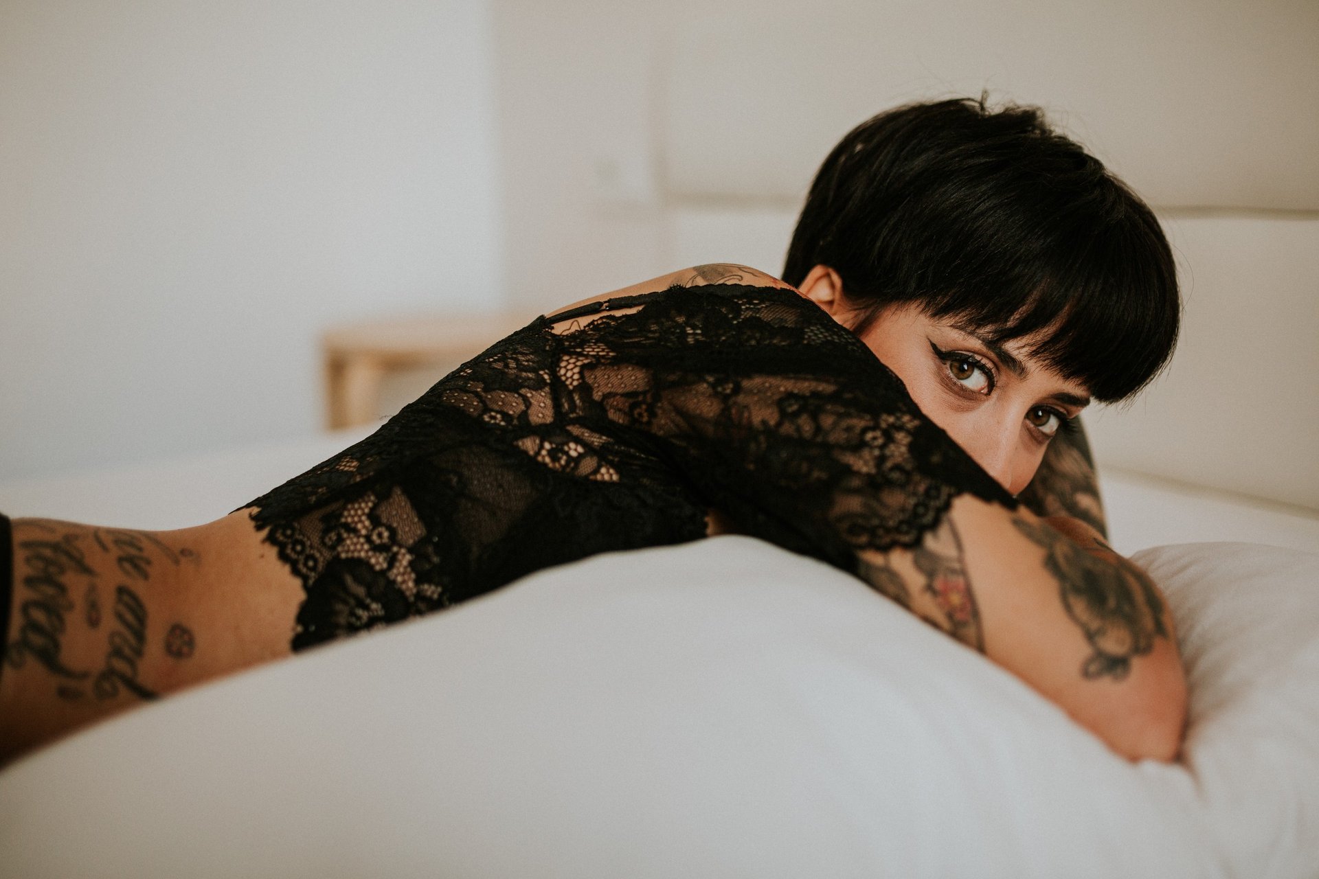 Mujer con cabello corto oscuro y tatuajes, recostada en una cama de sábanas blancas con un top de encaje negro, mirando a cámara con confianza. Luz natural suave. Fotografía de boudoir de Víctor Ruiz, Granada.
