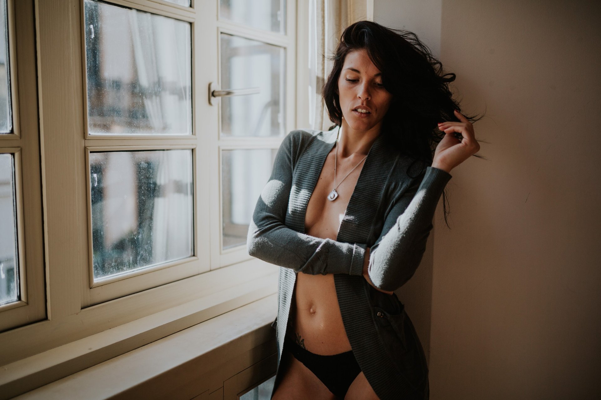 Mujer de cabello oscuro ondulado con una rebeca gris y ropa interior negra, apoyada en el alféizar de una ventana con luz natural lateral, una mano en el pelo y los ojos cerrados. Fotografía de boudoir de Víctor Ruiz, Granada.
