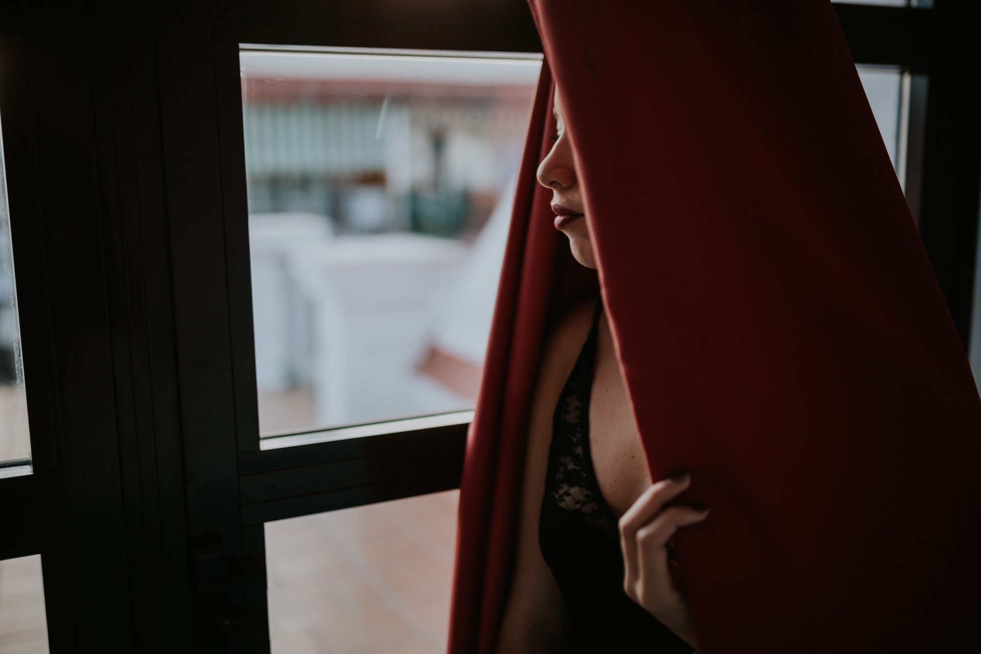 Mujer de perfil con lencería de encaje negro junto a una ventana, sujetando una cortina roja, iluminada por la luz exterior con alto contraste. Fotografía de boudoir de Víctor Ruiz, Granada.