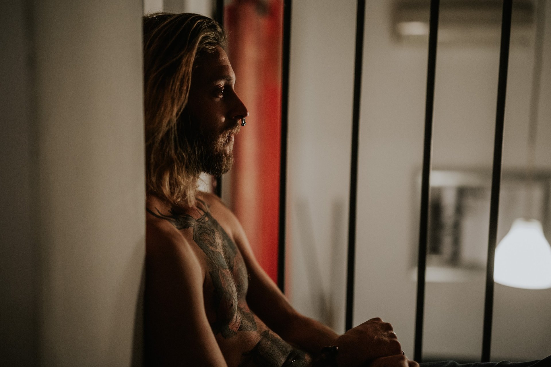 Hombre de pelo largo y barba sin camisa, con tatuajes, apoyado contra una pared con luz cálida, mirada pensativa. Fotografía de boudoir de Víctor Ruiz, Granada.