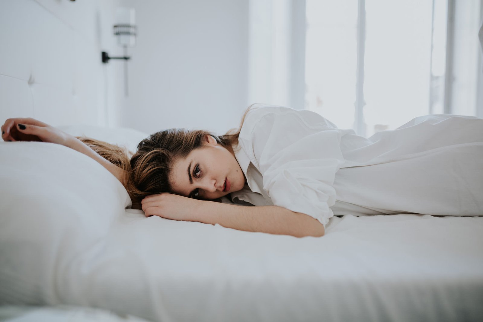 Mujer recostada boca abajo en una cama de sábanas blancas, apoyada en los codos y mirando a cámara con expresión serena, en una habitación luminosa con cortinas translúcidas. Fotografía de boudoir de Víctor Ruiz, Granada.