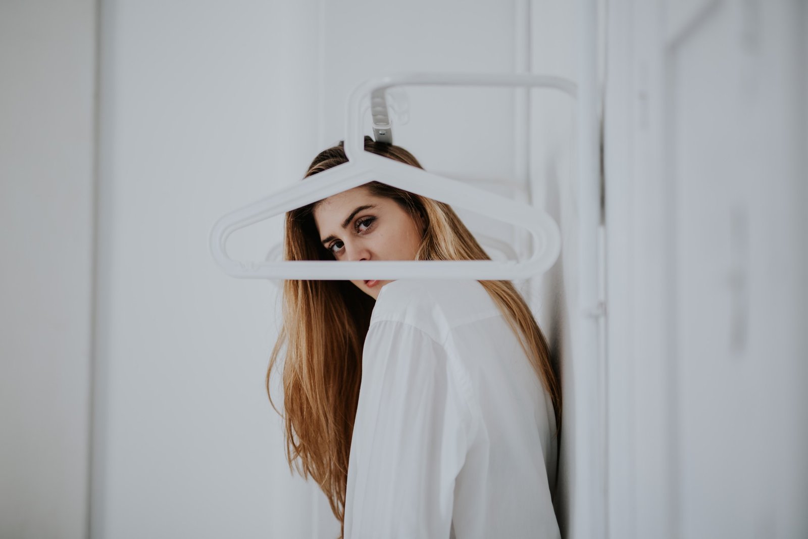 Primer plano de una mujer con camisa blanca mirando a cámara con una percha blanca delante del rostro, fondo blanco luminoso. Fotografía de boudoir de Víctor Ruiz, Granada.
