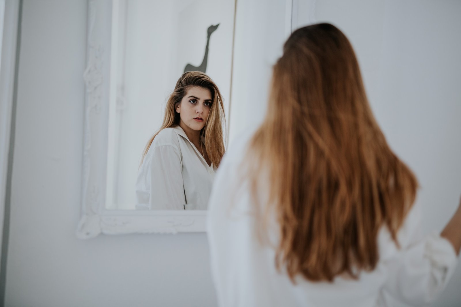 Mujer de espaldas con camisa blanca mirándose en un espejo de marco blanco, en una habitación luminosa y minimalista. Fotografía de boudoir de Víctor Ruiz, Granada.