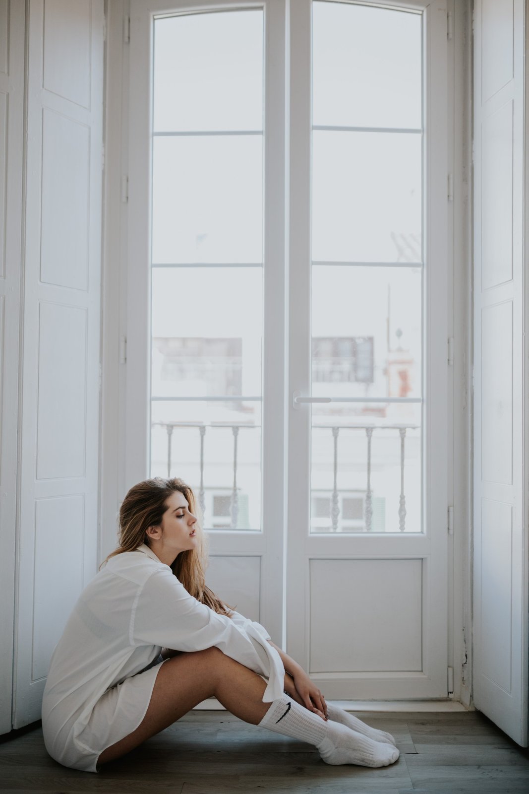 Mujer sentada en el suelo junto a una gran puerta arqueada, abrazando sus rodillas y mirando a un lado, con luz natural suave. Fotografía de boudoir de Víctor Ruiz, Granada.