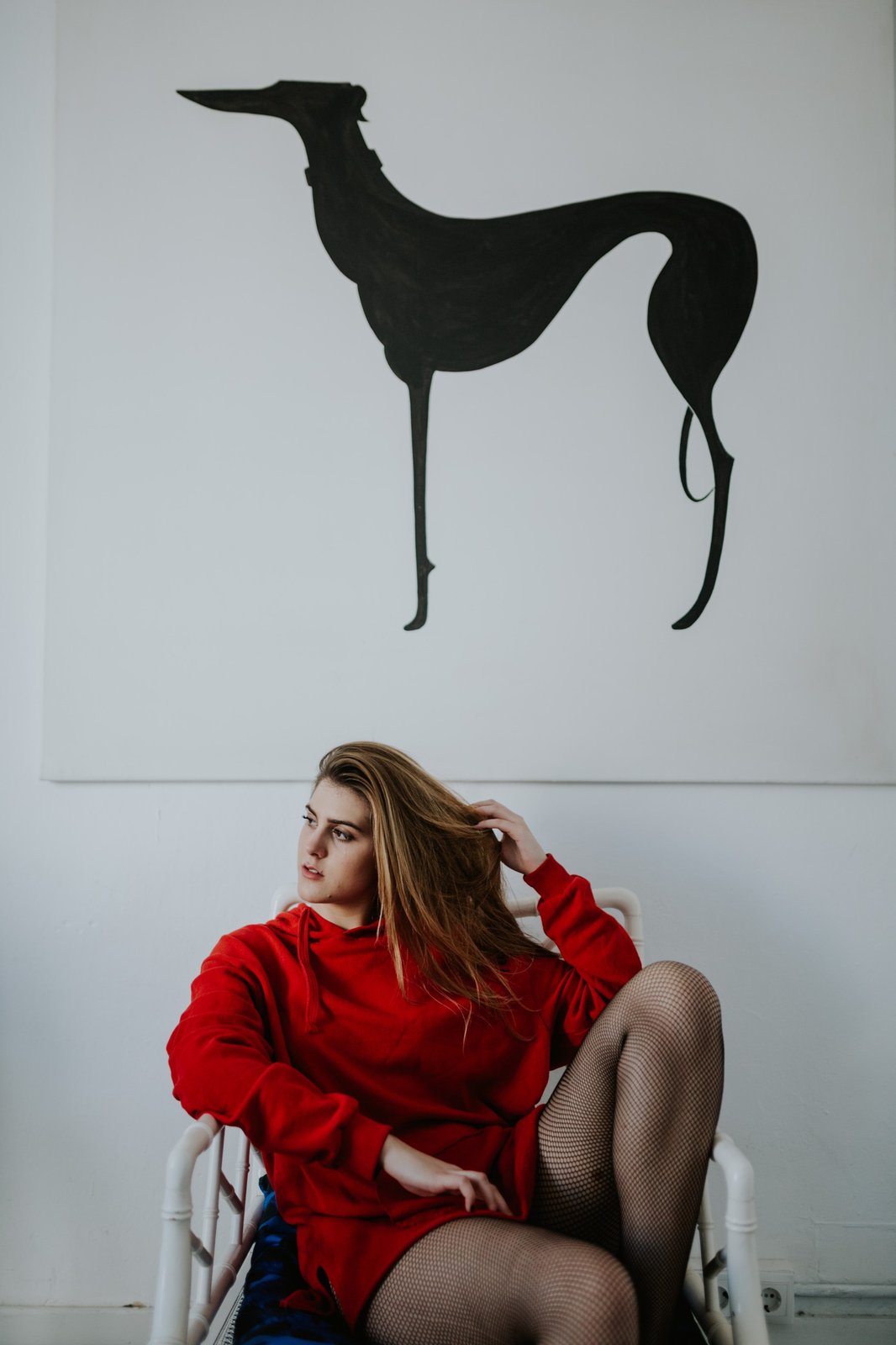 Mujer con sudadera roja oversized y medias de red sentada en una silla blanca frente a un cuadro con la silueta negra de un galgo. Fotografía de boudoir de Víctor Ruiz, Granada.