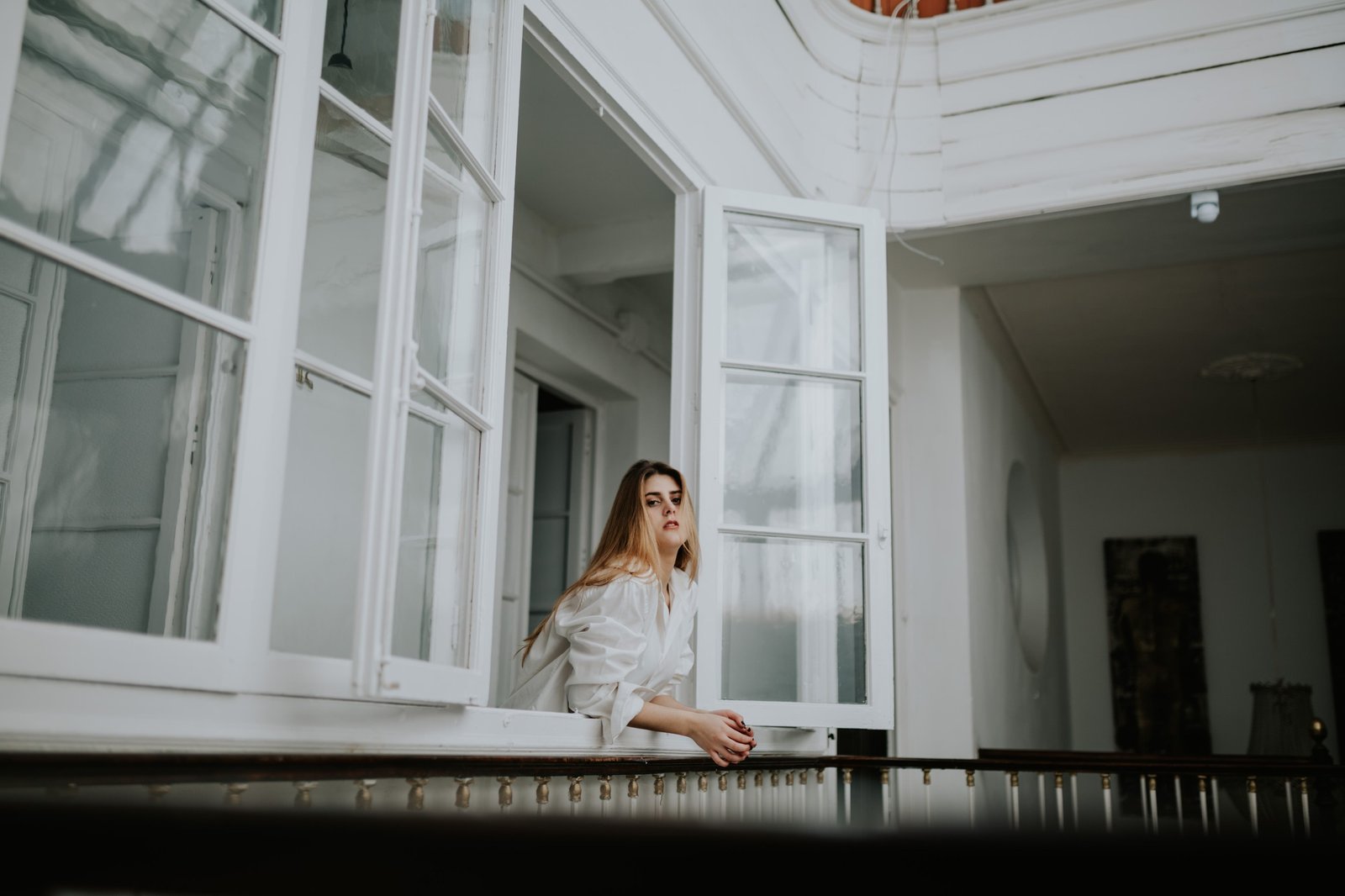 Mujer asomada a una ventana abierta con los antebrazos apoyados en el alféizar, camisa blanca oversized y luz natural suave. Fotografía de boudoir de Víctor Ruiz, Granada.