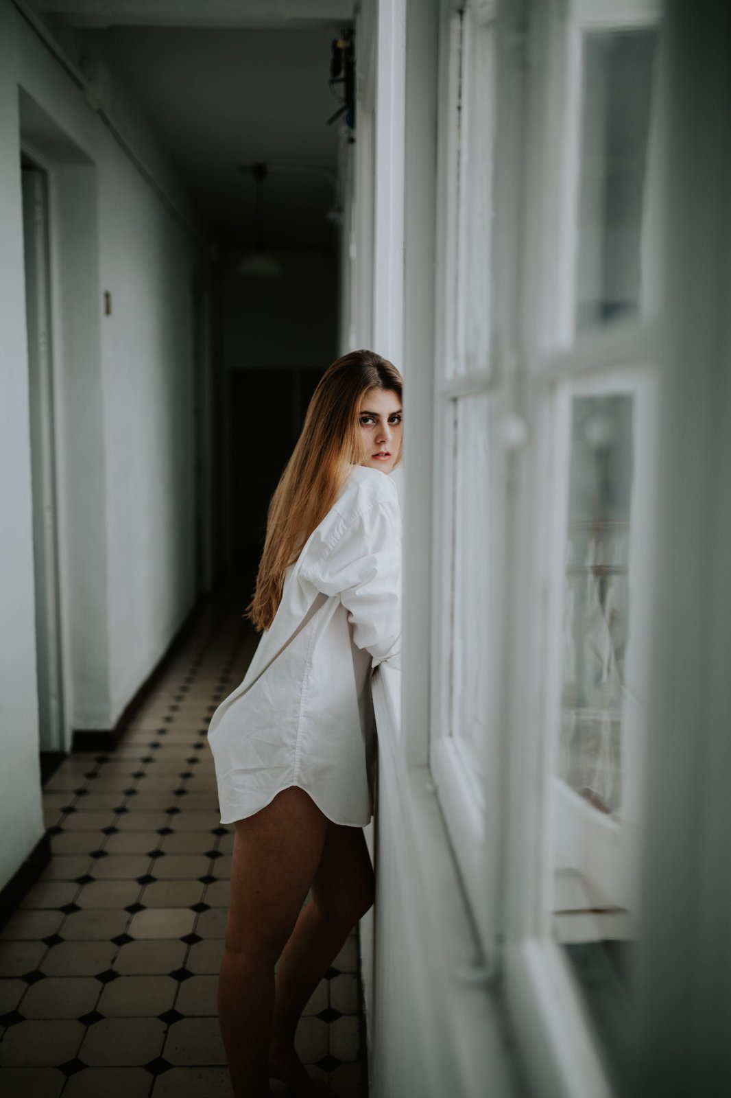 Mujer con camisa blanca oversized en un pasillo luminoso, mirando a cámara por encima del hombro junto a una ventana. Fotografía de boudoir de Víctor Ruiz, Granada.