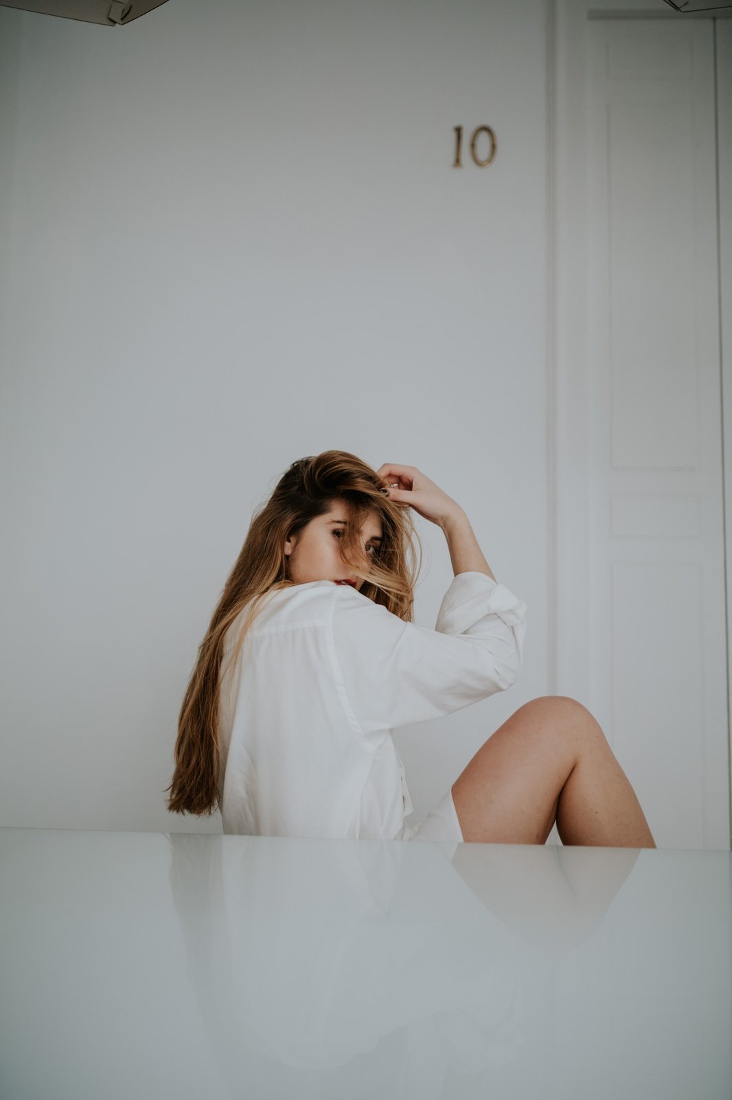 Mujer sentada en una superficie blanca reflectante mirando por encima del hombro con una mano en el pelo, en una habitación blanca y luminosa. Fotografía de boudoir de Víctor Ruiz, Granada.
