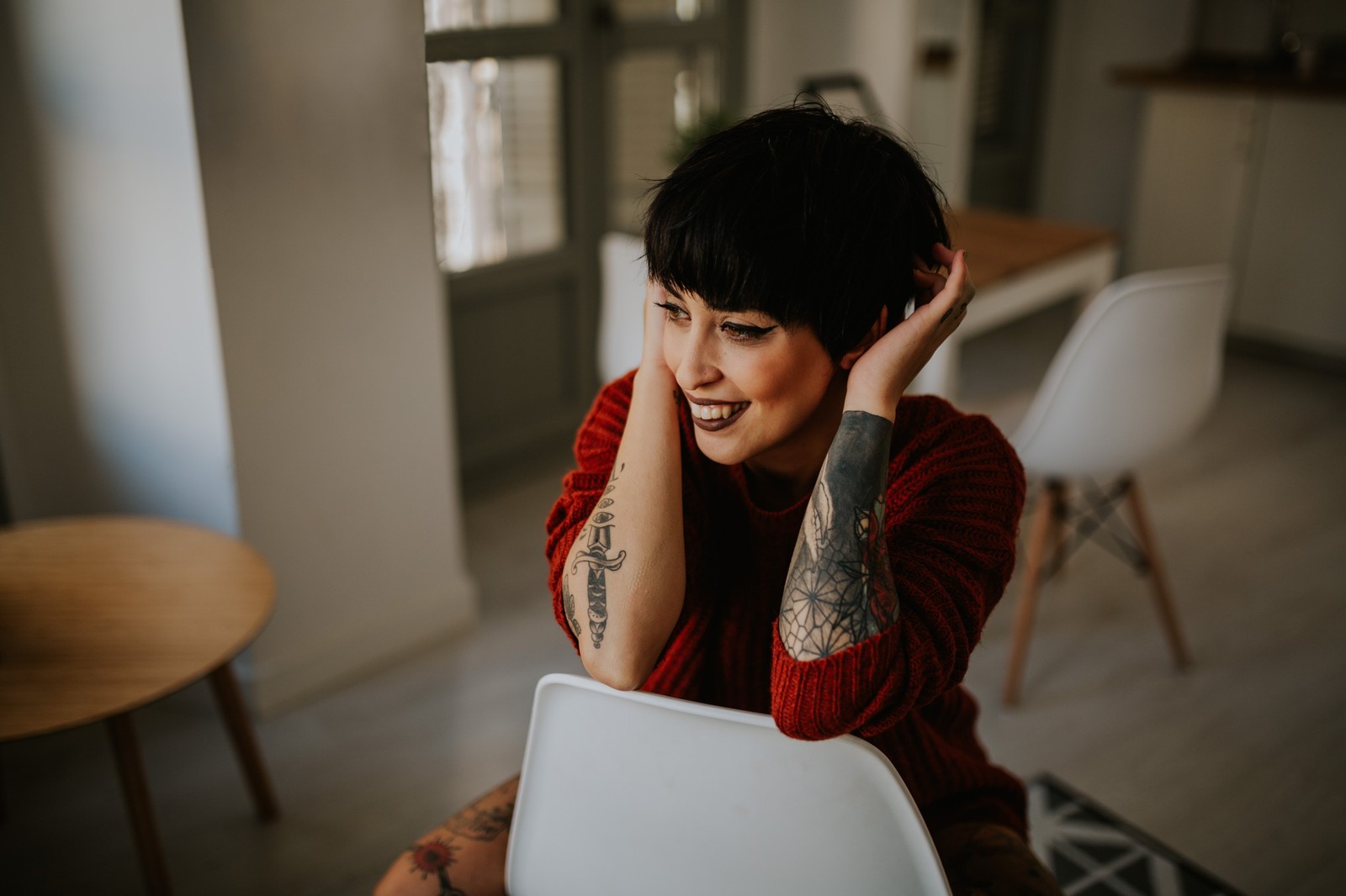 Mujer con cabello corto y tatuajes sentada en una silla blanca sonriendo y tocándose el pelo, suéter rojo de punto, interior luminoso. Fotografía de boudoir de Víctor Ruiz, Granada.