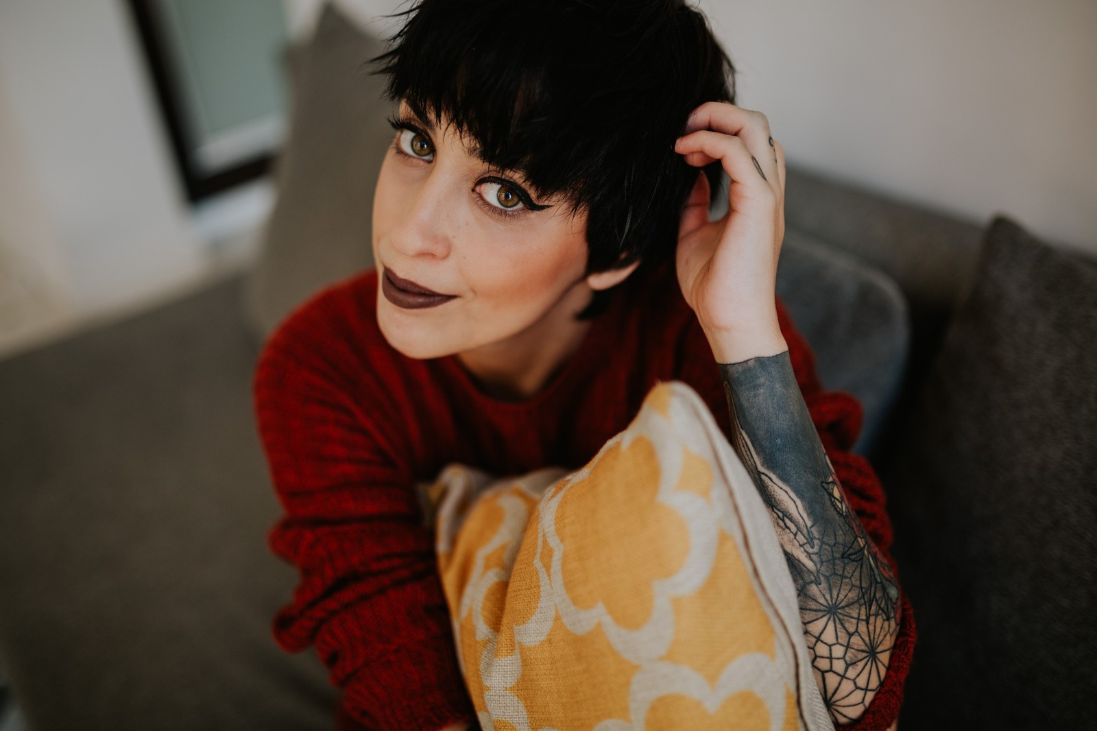 Primer plano de una mujer con tatuajes y suéter rojo de cuello alto, mano en el pelo, mirada intensa, luz suave. Fotografía de boudoir de Víctor Ruiz, Granada.