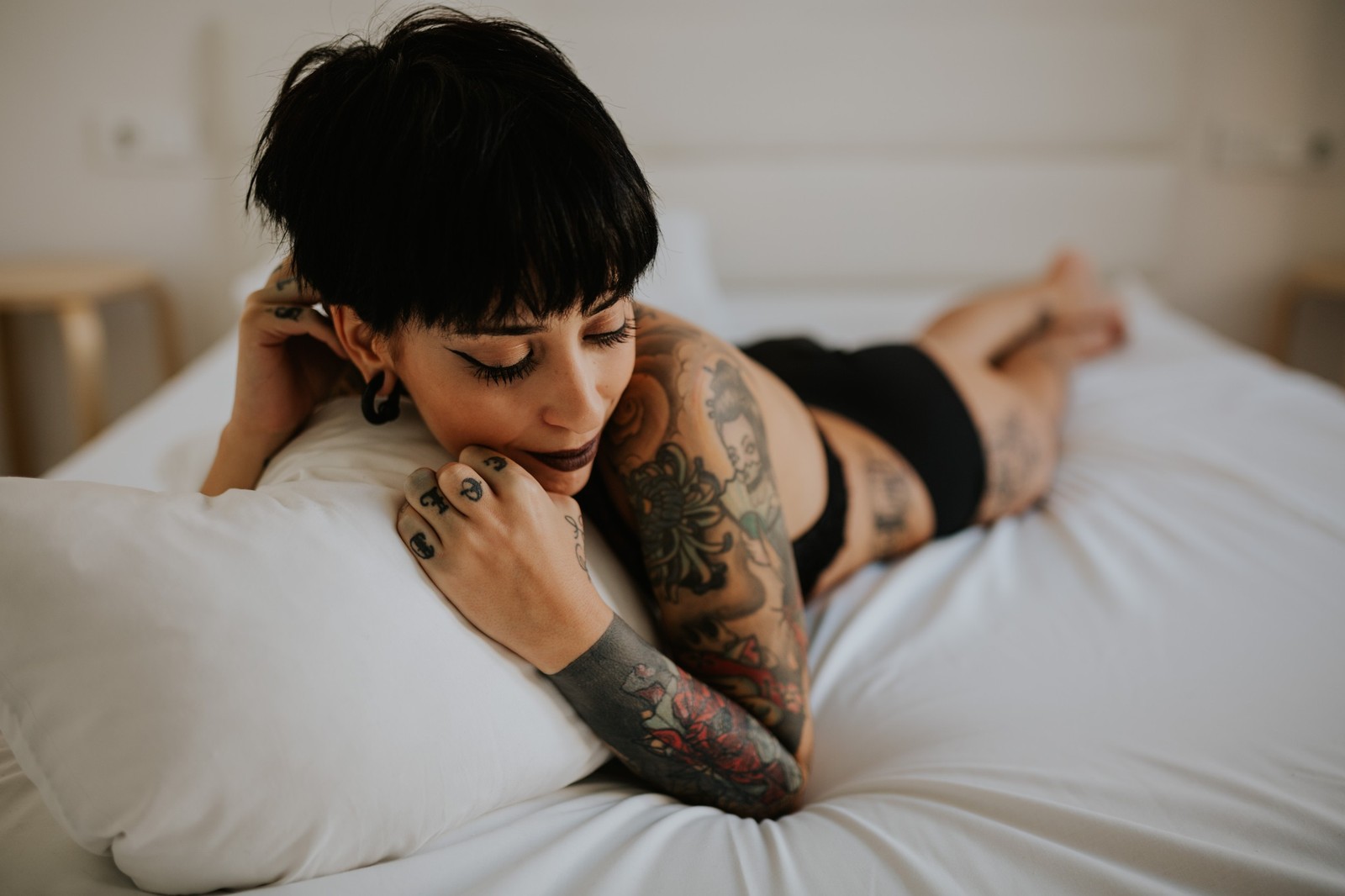 Mujer con tatuajes tumbada boca abajo en una cama blanca con ropa interior negra, descansando la cabeza en las manos, luz natural serena. Fotografía de boudoir de Víctor Ruiz, Granada.