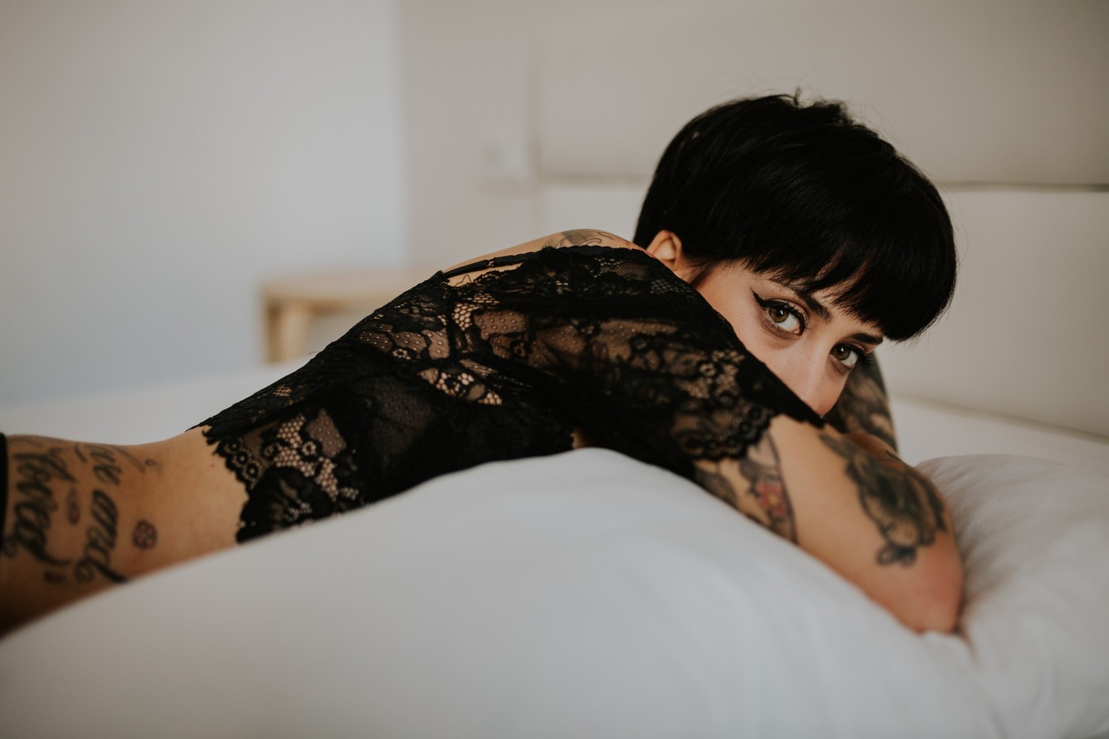 Mujer con cabello corto y tatuajes en un top de encaje negro sobre una cama blanca, mirando a cámara con calma, luz difusa. Fotografía de boudoir de Víctor Ruiz, Granada.