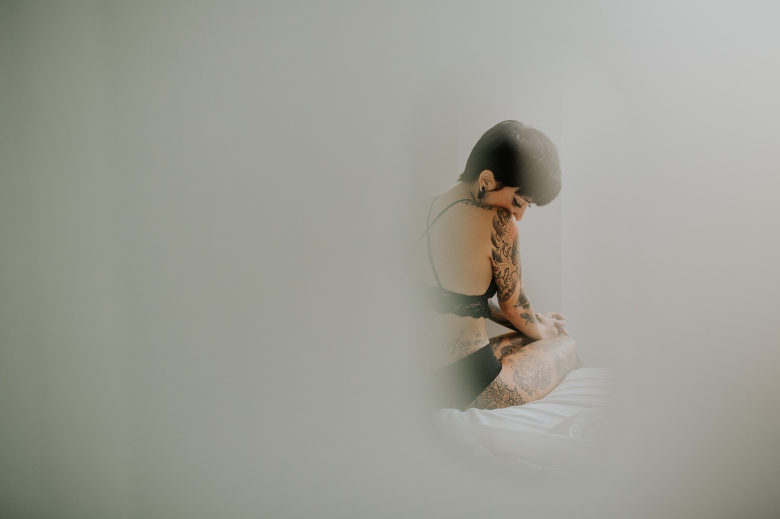 Mujer con tatuajes sentada en una cama con lencería negra en pose reflexiva, luz suave y difusa, ambiente etéreo. Fotografía de boudoir de Víctor Ruiz, Granada.