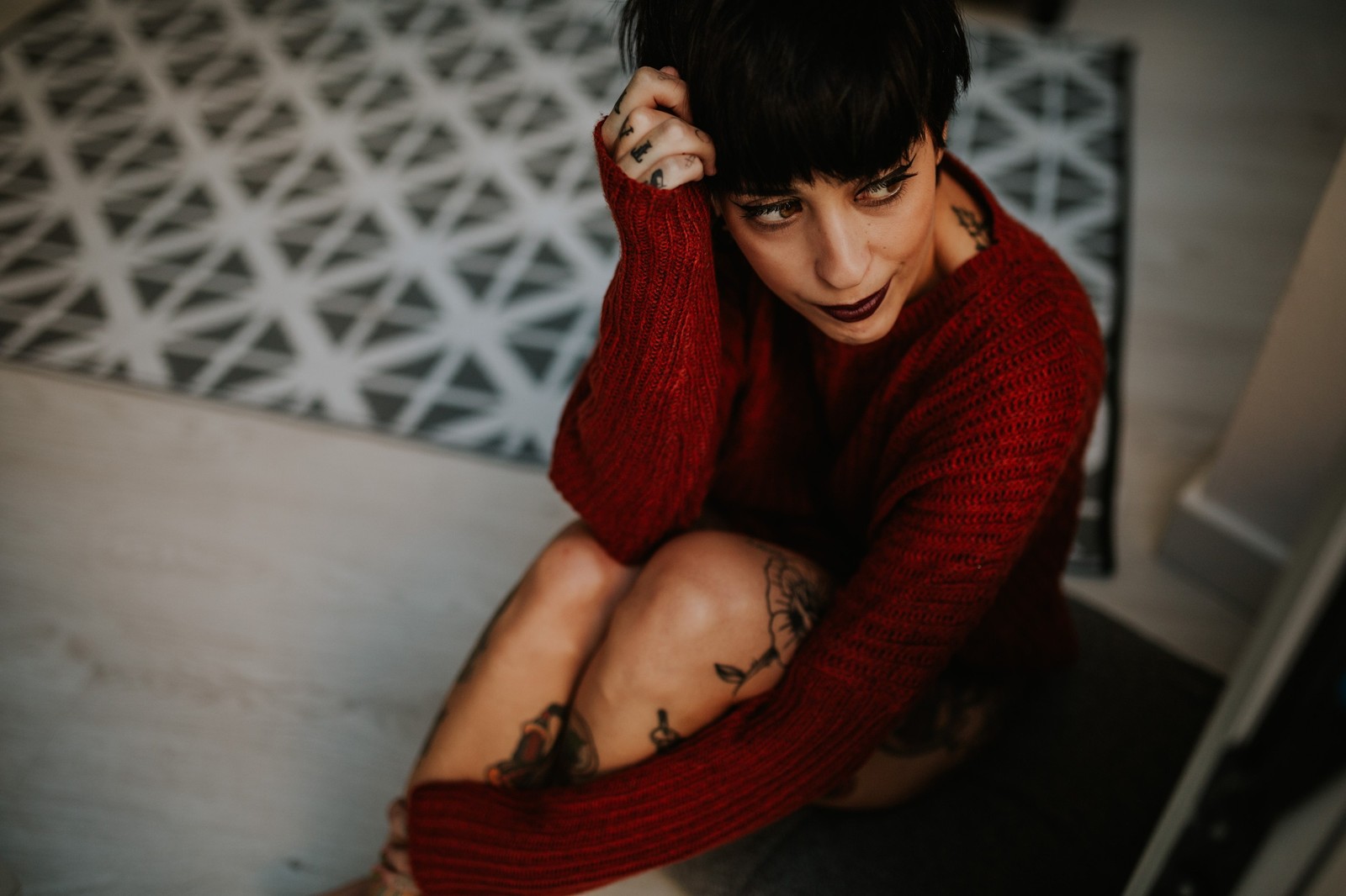 Mujer joven con cabello corto y tatuajes sentada en el suelo con un suéter rojo, mano en la cabeza, luz natural que crea un ambiente íntimo y acogedor. Fotografía de boudoir de Víctor Ruiz, Granada.